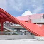 Low-angle view of a red mesh-covered roof swooping down to the platform level near an outdoor staircase and observation platform.