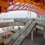 Architectural interior showing a glass-walled staircase connecting two levels while a sightseeing train moves past on the tracks below.