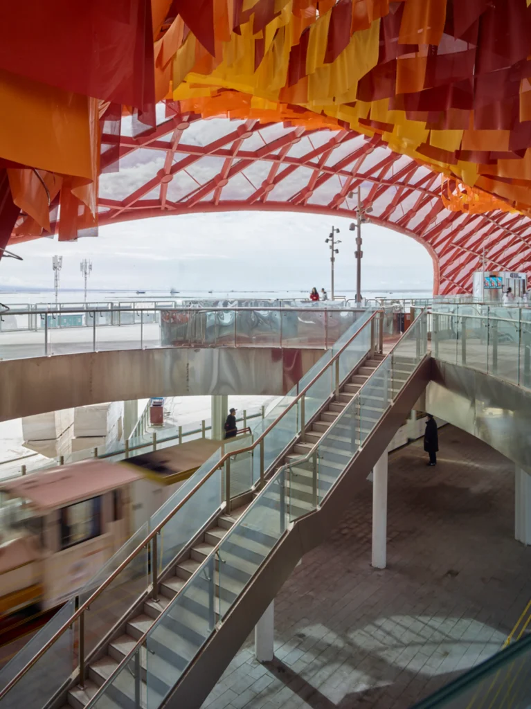 Architectural interior showing a glass-walled staircase connecting two levels while a sightseeing train moves past on the tracks below.