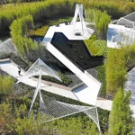 Aerial view of white geometric steel structures, curved paths and a dark reflecting pool set among dense bamboo