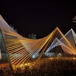 Night close-up of curved and angular white steel frameworks glowing under warm artificial light