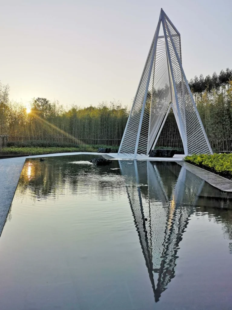Sunset view of a tall white pyramidal steel structure reflected in a still rectangular pool