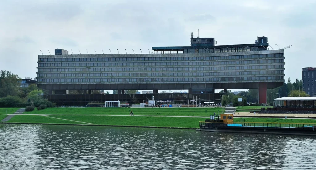 Long elevated concrete hotel building with continuous horizontal window bands, raised on massive piers above a green riverside park