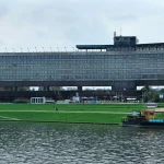 Long elevated concrete hotel building with continuous horizontal window bands, raised on massive piers above a green riverside park