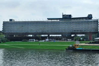 Long elevated concrete hotel building with continuous horizontal window bands, raised on massive piers above a green riverside park
