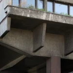 Close-up of weathered concrete underside showing deep angular beams, recessed voids and vegetation growing along the upper edge of the elevated structure