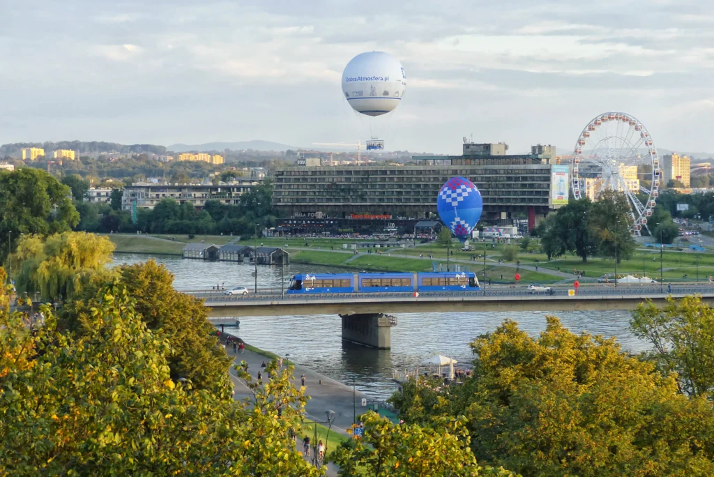 Distant view of Forum Hotel across the river with hot-air balloons, a Ferris wheel and a blue tram crossing a bridge in the foreground