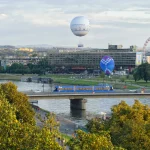 Distant view of Hotel Forum across the river with hot-air balloons, a Ferris wheel and a blue tram crossing a bridge in the foreground