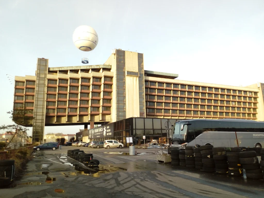 Street-level view of the hotel’s stepped end elevation and long façade, with coaches, parked cars and a large observation balloon overhead