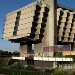 Angular concrete wing of the hotel raised on thick piers, with commercial signage and landscaping at ground level under a clear blue sky
