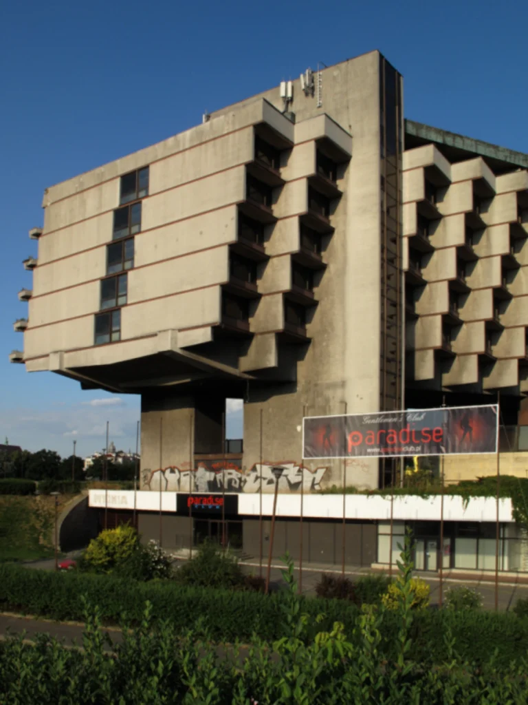 Angular concrete wing of the hotel raised on thick piers, with commercial signage and landscaping at ground level under a clear blue sky
