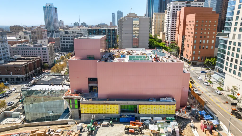 Frank Gehry’s Pink Colburn School Expansion to Open in Los Angeles in 2027_ Aerial view of the pink-clad Colburn School expansion under construction amid downtown Los Angeles skyscrapers