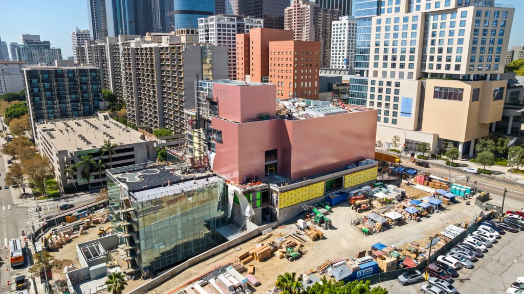 Wider aerial showing the pink building volumes, glass section, and active construction site with surrounding towers