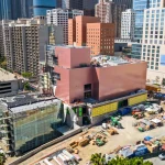 Wider aerial showing the pink building volumes, glass section, and active construction site with surrounding towers
