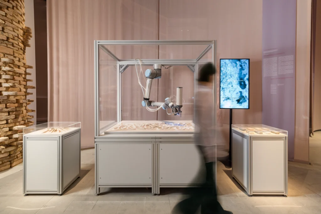 Exhibition view of a robotic biocementation setup inside a glass case with a person walking past and display screens