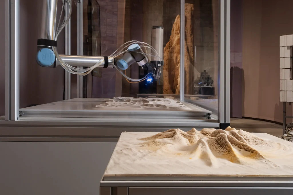 Robotic arm inside a glass enclosure depositing material onto a textured sand landscape model
