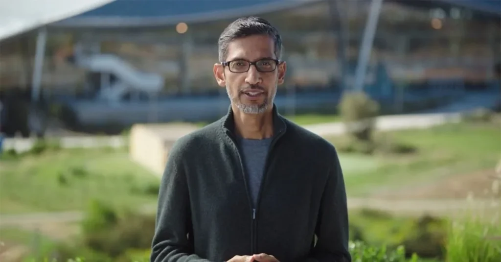 Sundar Pichai standing outdoors in a dark sweater, speaking toward the camera with Google campus buildings behind him