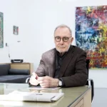 Gerhard Richter, an elderly man with short grey hair, a grey beard, and round black-rimmed glasses, sits at a glass-topped desk with his hands clasped. He wears a brown textured blazer over a black shirt and a wristwatch. Behind him is a large colorful abstract painting; other abstract works hang on the white walls of a modern gallery or studio space with a grey sofa and bookshelf.