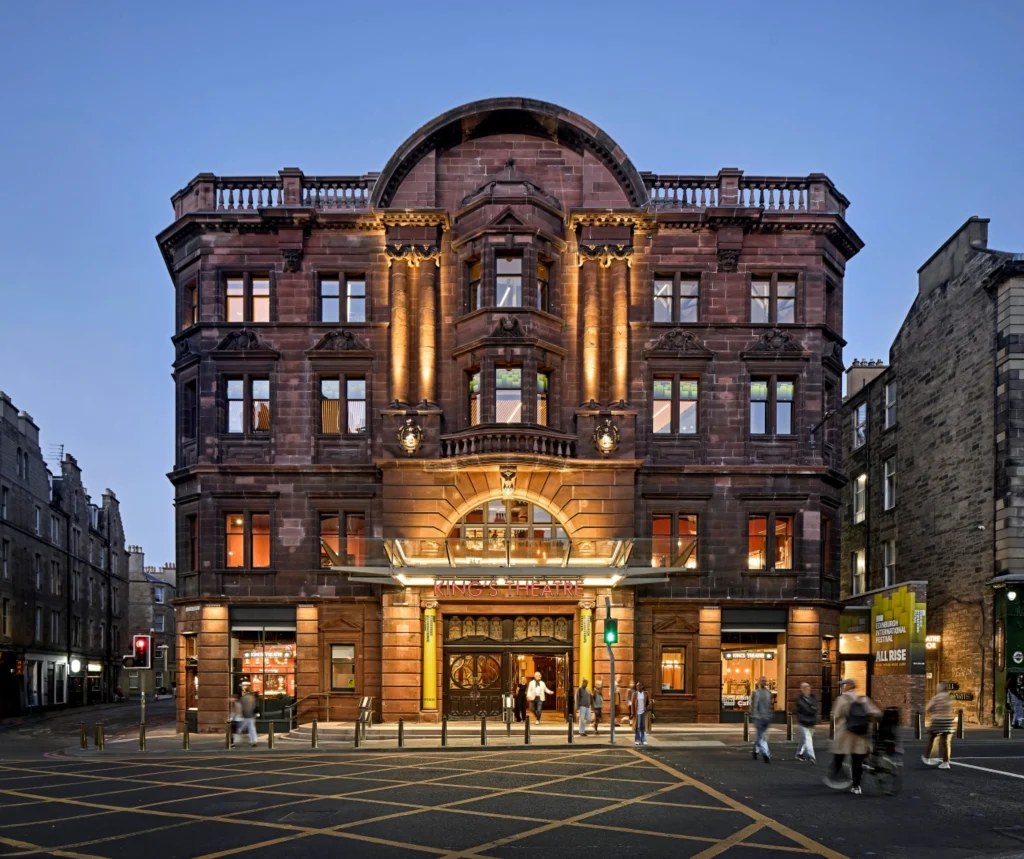 Night exterior of the illuminated red-stone King’s Theatre entrance with people walking past