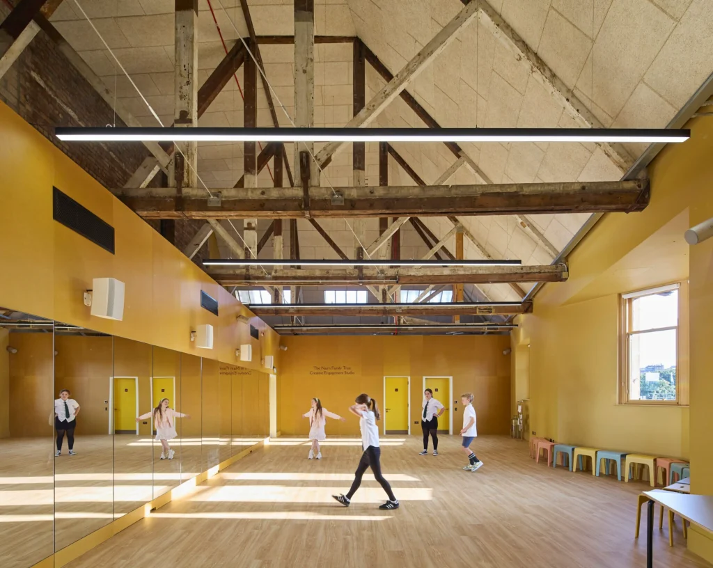 Children and adults moving in a bright yellow studio space with mirrored walls and timber roof structure