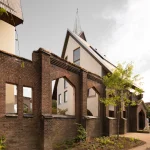 Weathered brick arches of the ruined church frame a view of the new cream-coloured houses with wooden roofs