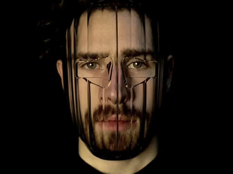 Close-up portrait of a bearded man wearing a transparent mask that vertically distorts and fragments his facial features against a black background