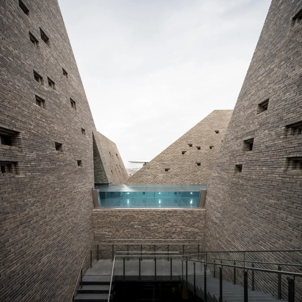 View looking up between tall angled brick towers toward an elevated outdoor pool with glass edge, under an overcast sky