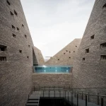 View looking up between tall angled brick towers toward an elevated outdoor pool with glass edge, under an overcast sky