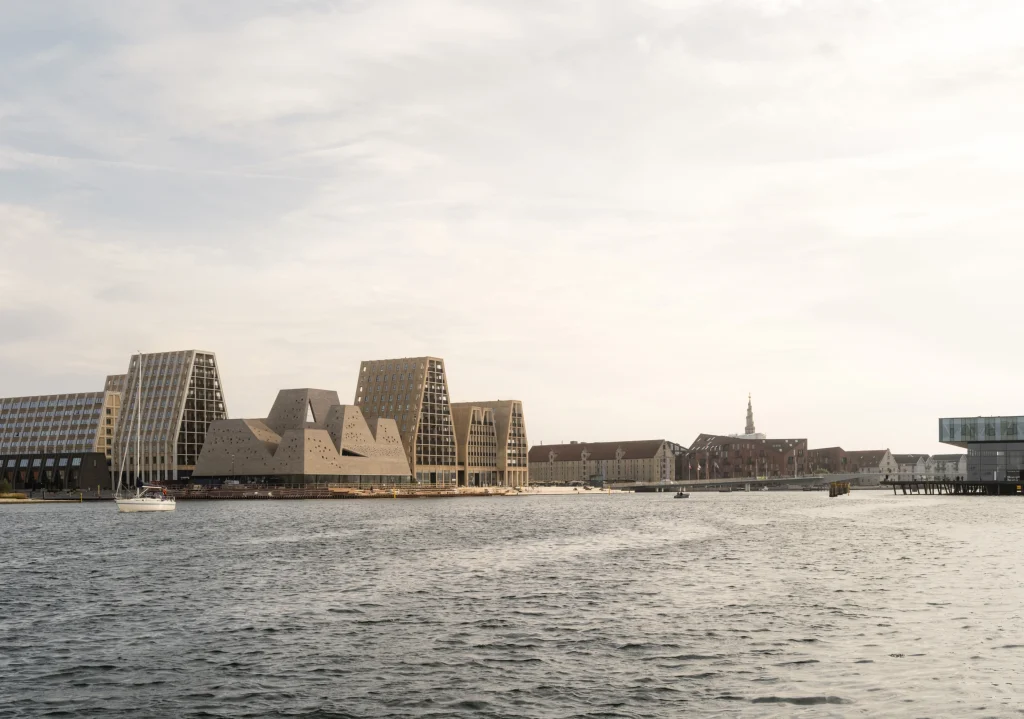 Wide harbour view of the Kengo Kuma's KAI building with its cluster of irregular brick volumes, surrounded by modern residential towers and historic cityscape