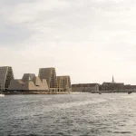 Wide harbour view of the KAI building with its cluster of irregular brick volumes, surrounded by modern residential towers and historic cityscape