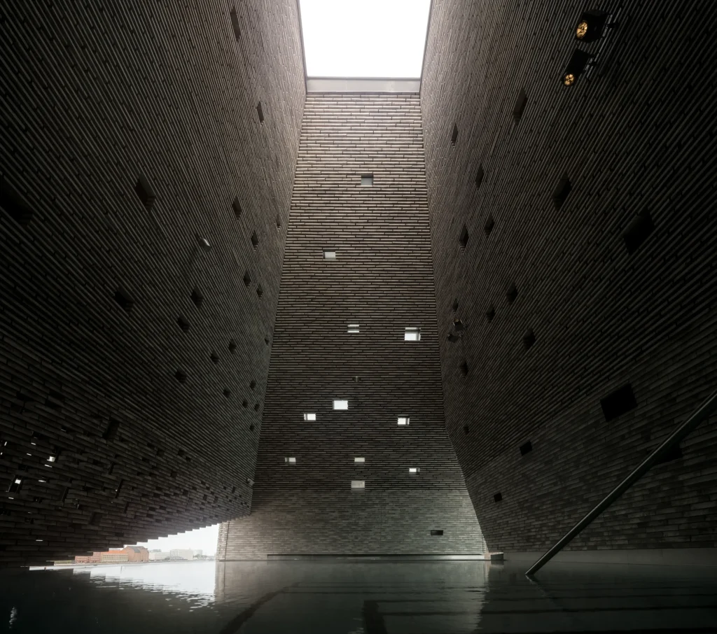 Dramatic upward interior view of a tall brick-lined shaft with scattered square openings and a reflective pool at the base