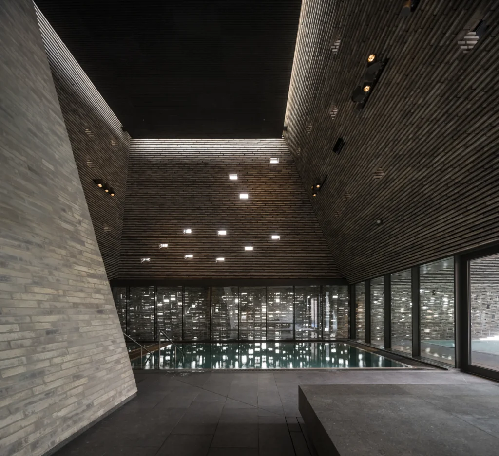 Indoor swimming pool under a high angled brick ceiling with recessed lights and large glass walls overlooking the exterior