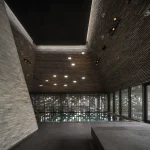 Indoor swimming pool under a high angled brick ceiling with recessed lights and large glass walls overlooking the exterior