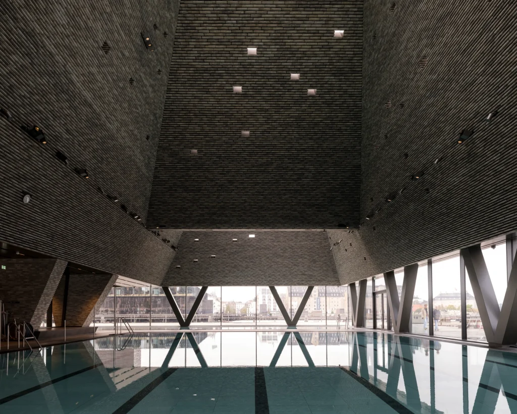 Long indoor pool reflecting the geometric brick ceiling and large V-shaped structural supports, with city views beyond the glass