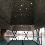 People swimming in an outdoor pool channelled between tall, textured brick walls under a clear blue sky