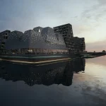 Twilight view of the illuminated KAI building reflected in calm harbour water, with surrounding architecture and a soft sunset