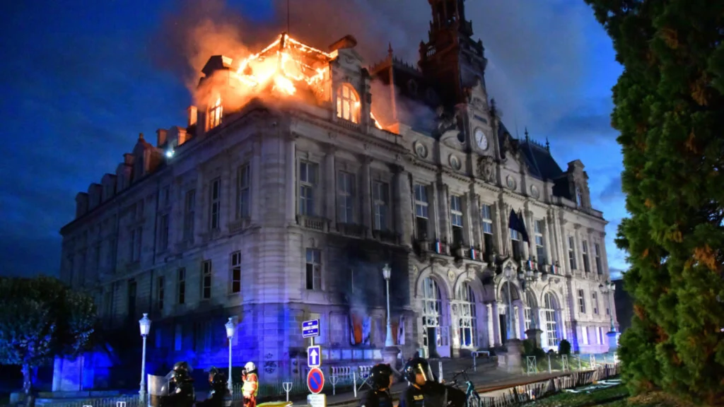 Side view of the burning town hall at dusk with police officers in the foreground and flames visible on the upper levels