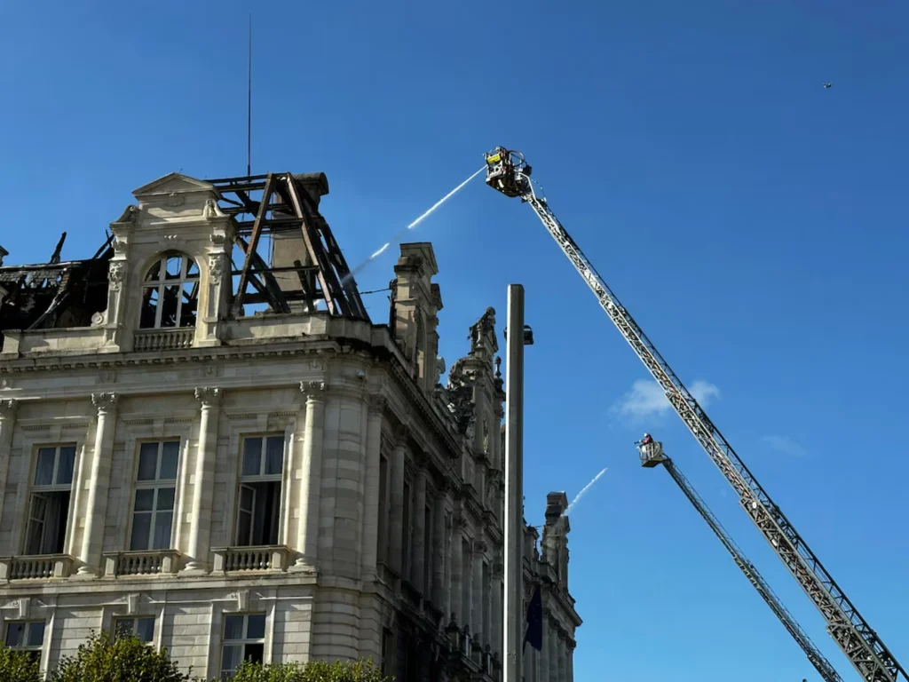Firefighters in elevated platforms spraying water onto the damaged roof of the Limoges town hall under a clear blue sky