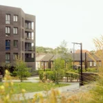 View through planting toward one of the dark brick pavilion blocks with balconies and street lighting