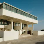 Side perspective of the elevated white house showing the long ribbon of green windows, concrete columns and beach visible in the distance