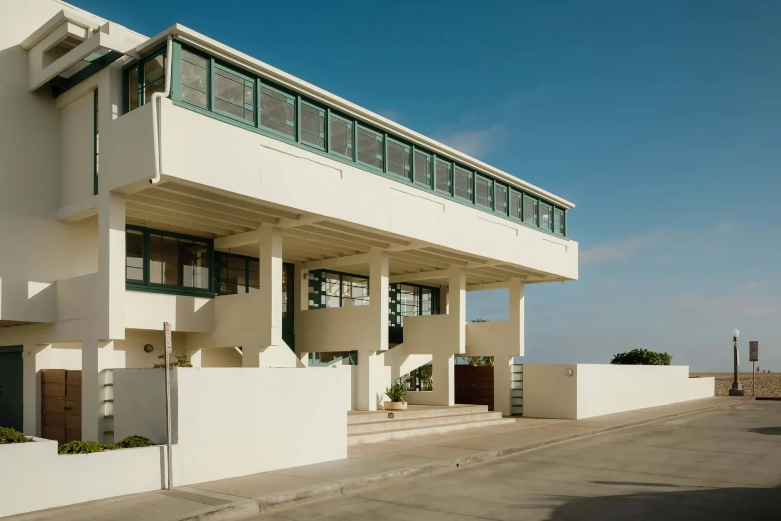 Side perspective of the elevated white house showing the long ribbon of green windows, concrete columns and beach visible in the distance