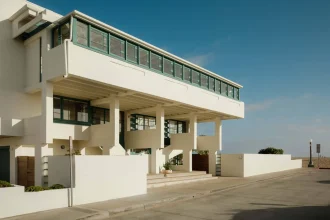 Side perspective of the elevated white house showing the long ribbon of green windows, concrete columns and beach visible in the distance