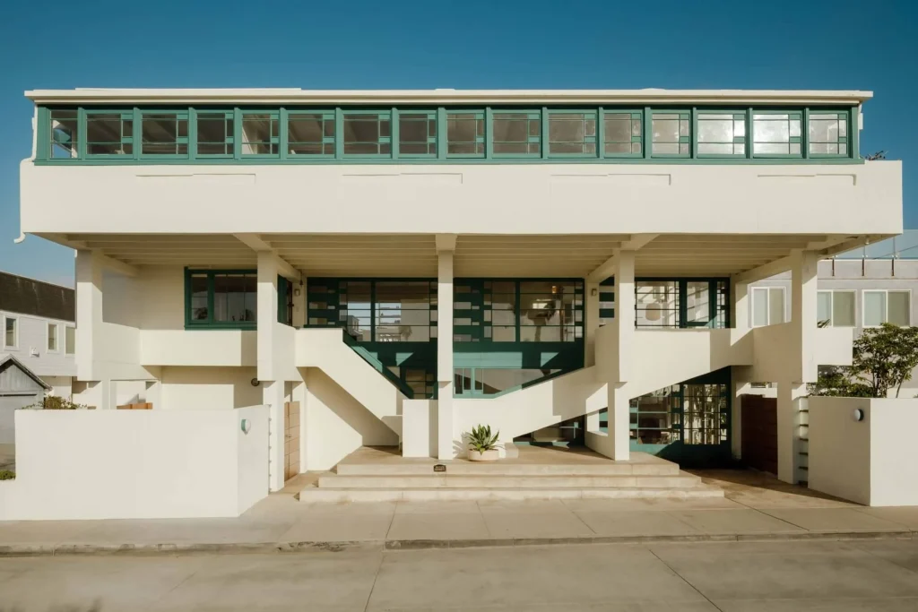 Front elevation of the white concrete house raised on pilotis with continuous green-framed windows along the upper level and open stairs
