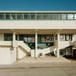 Front elevation of the white concrete house raised on pilotis with continuous green-framed windows along the upper level and open stairs
