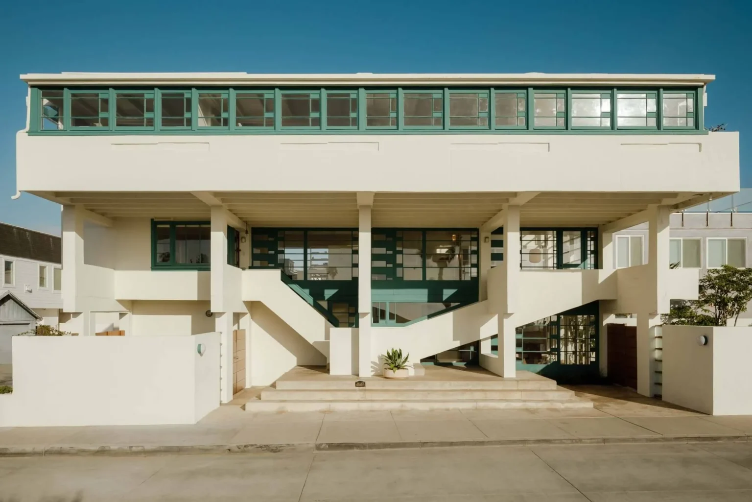 Front elevation of the white concrete house raised on pilotis with continuous green-framed windows along the upper level and open stairs