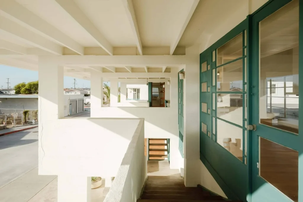 Interior view under the raised structure showing white concrete beams, open stairs and teal-framed glass doors leading deeper into the house