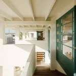 Interior view under the raised structure showing white concrete beams, open stairs and teal-framed glass doors leading deeper into the house