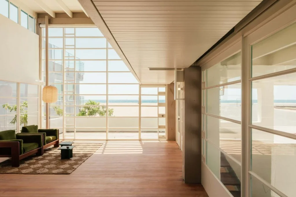 Bright interior living area with floor-to-ceiling grid windows overlooking the beach, green armchairs and wooden floors