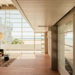 Bright interior living area with floor-to-ceiling grid windows overlooking the beach, green armchairs and wooden floors