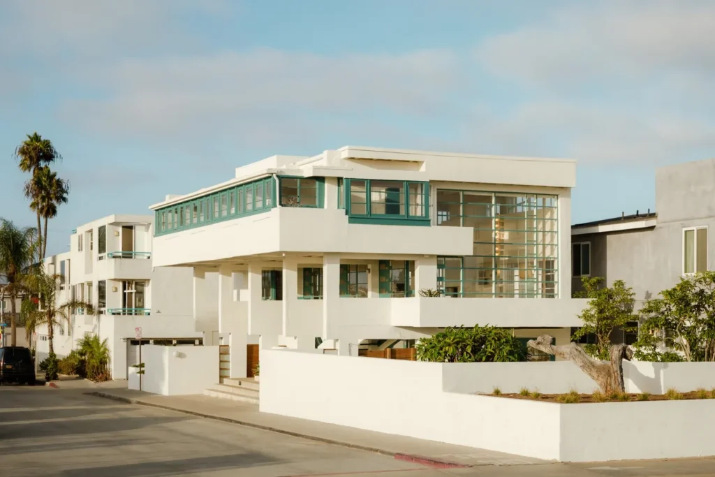 Exterior corner view of the white modernist house with green window frames, surrounding low white walls and neighbouring houses